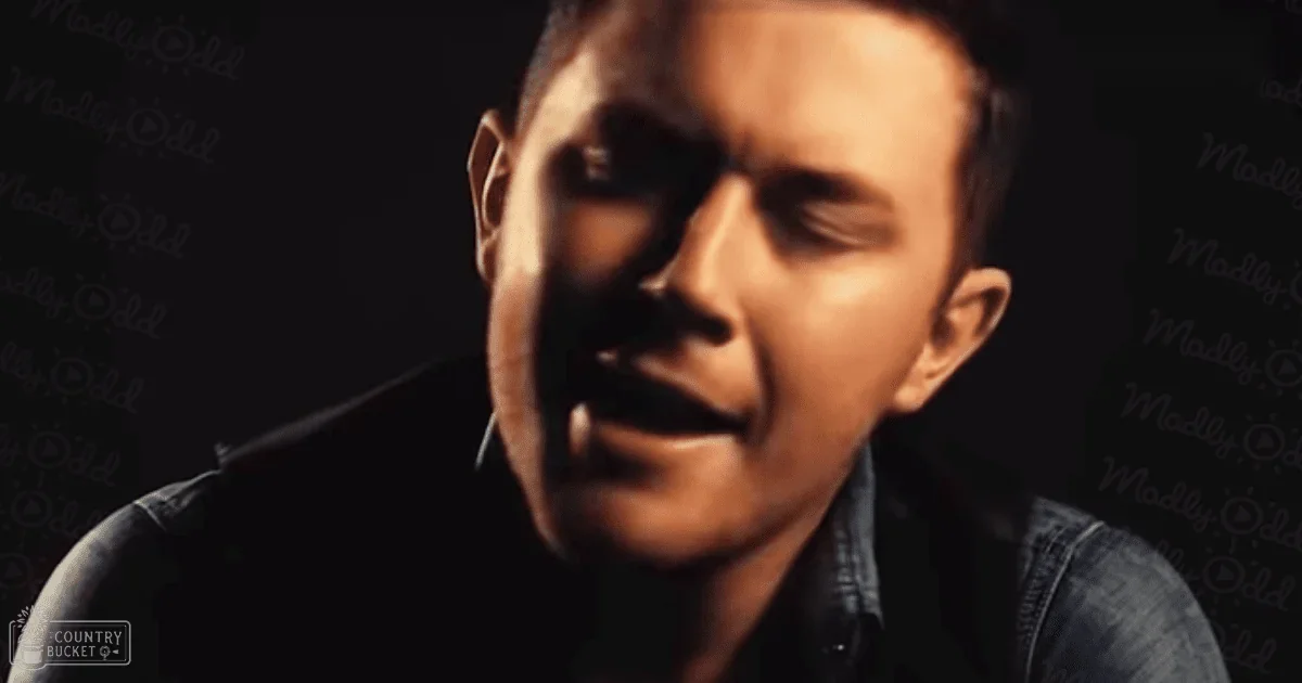 A young adult male stands indoors, wearing a relaxed blue denim shirt or jacket with subtle fading and a collared neckline. The dark textured background suggests an indoor setting with artificial lighting.