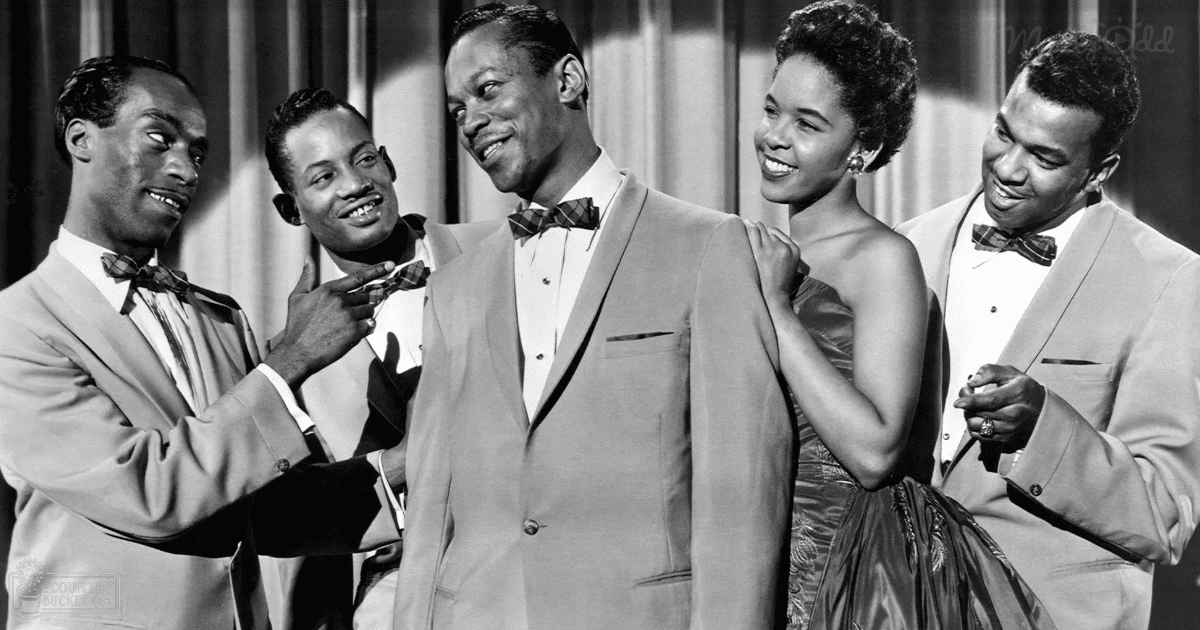 A group of performers in a studio setting with curtains and professional lighting, dressed formally in light-colored suits and a satin dress.