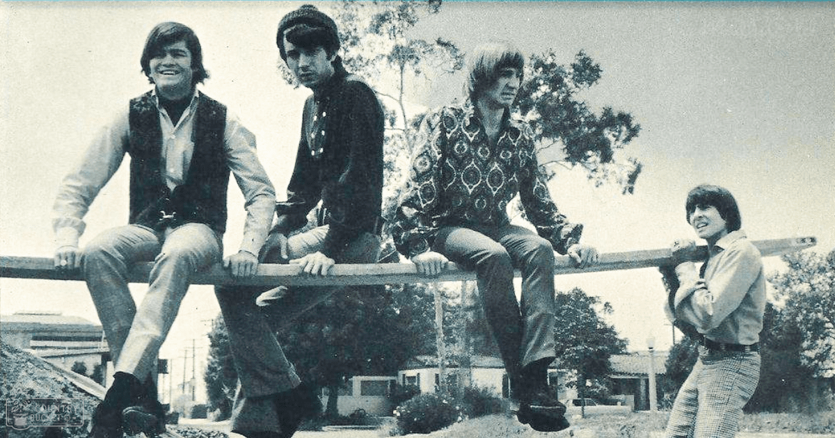 Four young adults standing outdoors, wearing casual spring/summer attire with trees and buildings in the background; daytime lighting; black and white image.