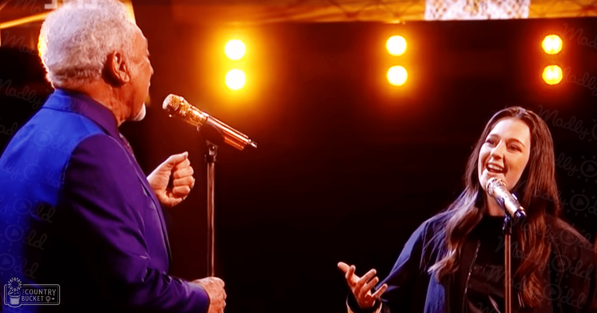 A duet performance scene with two adult performers on a stage illuminated by bright orange lights, wearing dark blue suits and black inner garments, set against a dark background.