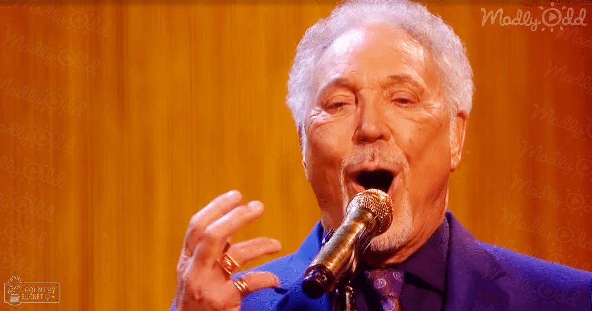 A middle-aged male singer in a formal blue suit with a purple circular-patterned tie, performing on stage at what seems to be a concert or performance setting.