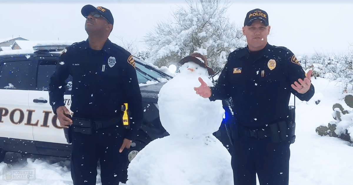 Two police officers stand outdoors in winter, with a snowman in front of them. Snow covers the ground and trees behind a partially visible police vehicle.