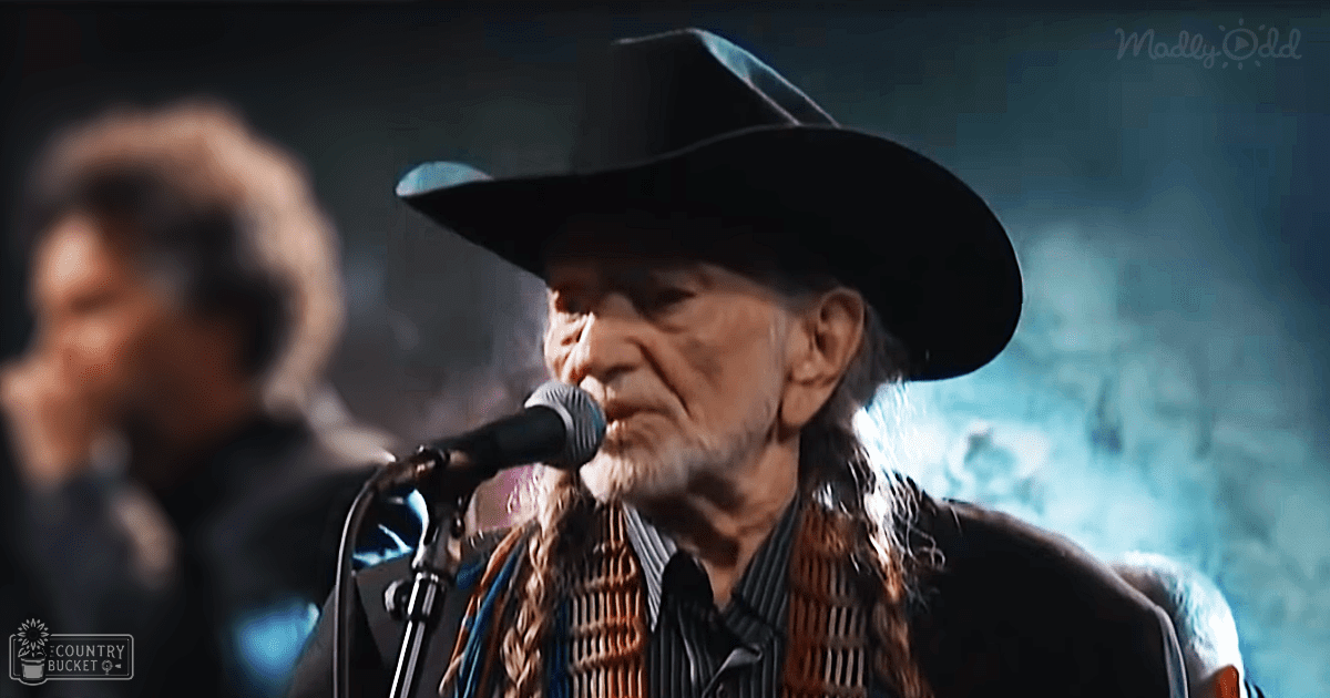 A middle-aged man wearing a cowboy hat and patterned scarf, standing on stage at night during what appears to be a concert or performance with dim lighting focused on him.