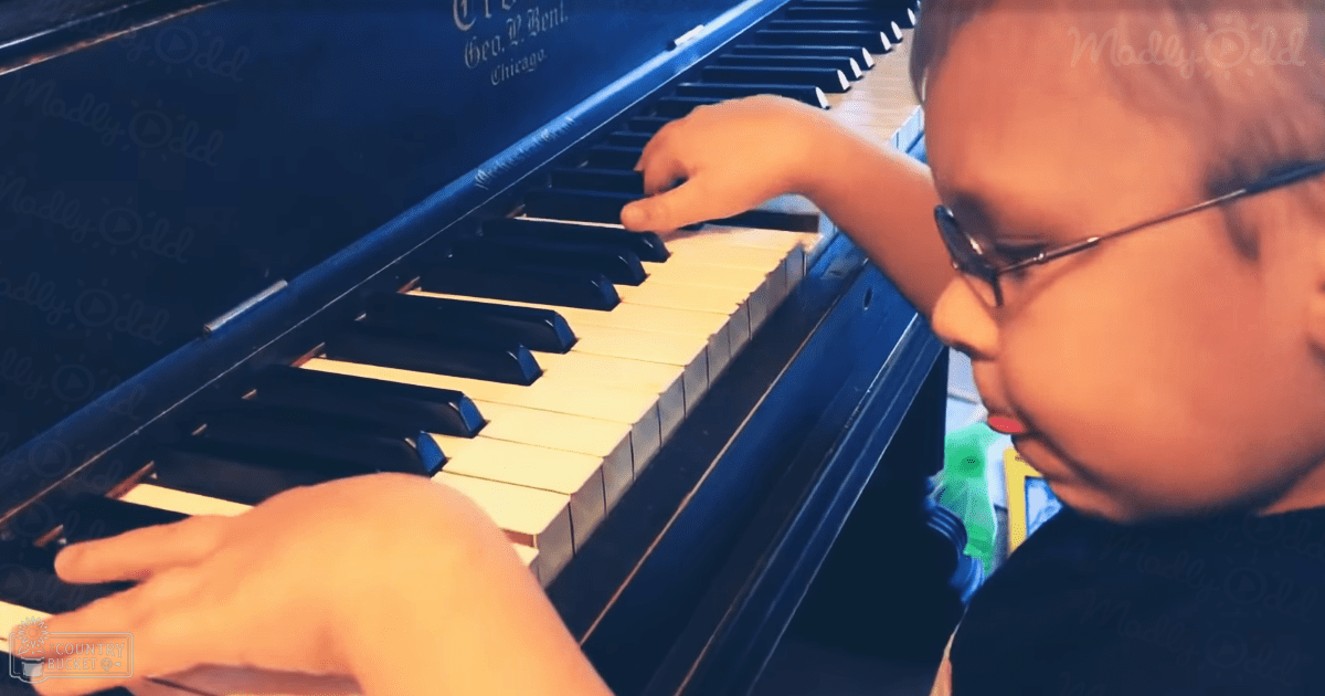 A young male child plays a dark blue piano in an indoor home setting, wearing a dark-colored shirt with short sleeves and a crew neckline.