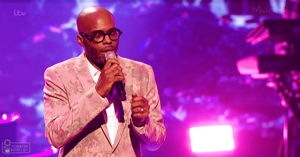 An adult male singer in a patterned blazer at a live performance setting under stage lighting.