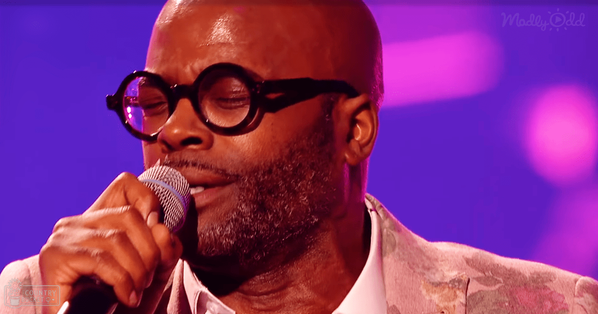Adult male singer in a patterned blazer with floral designs at a music event, microphone and purple stage lighting visible.