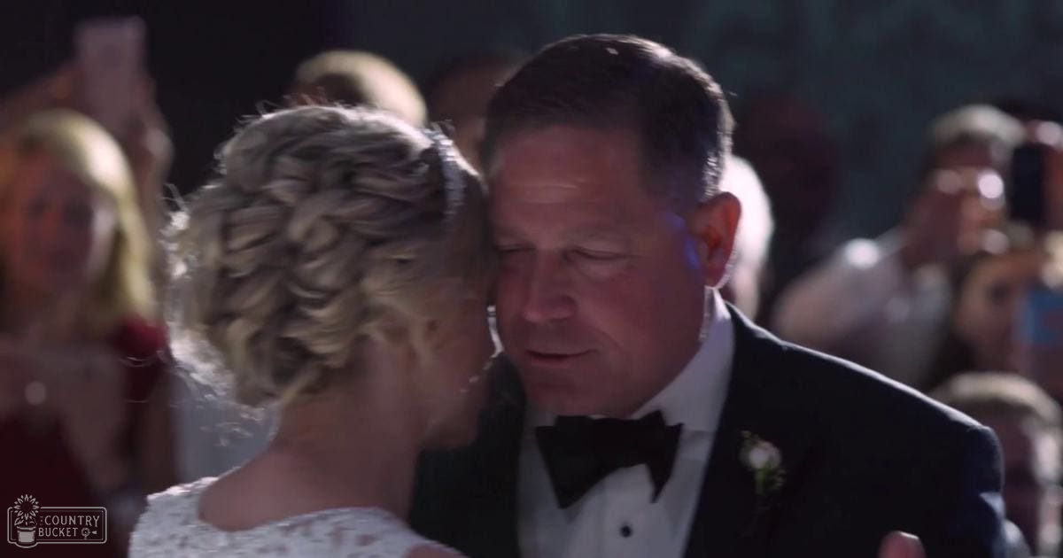 A young adult female in a white lace wedding dress dances with an older male wearing a black tuxedo at an indoor evening wedding reception, surrounded by guests.