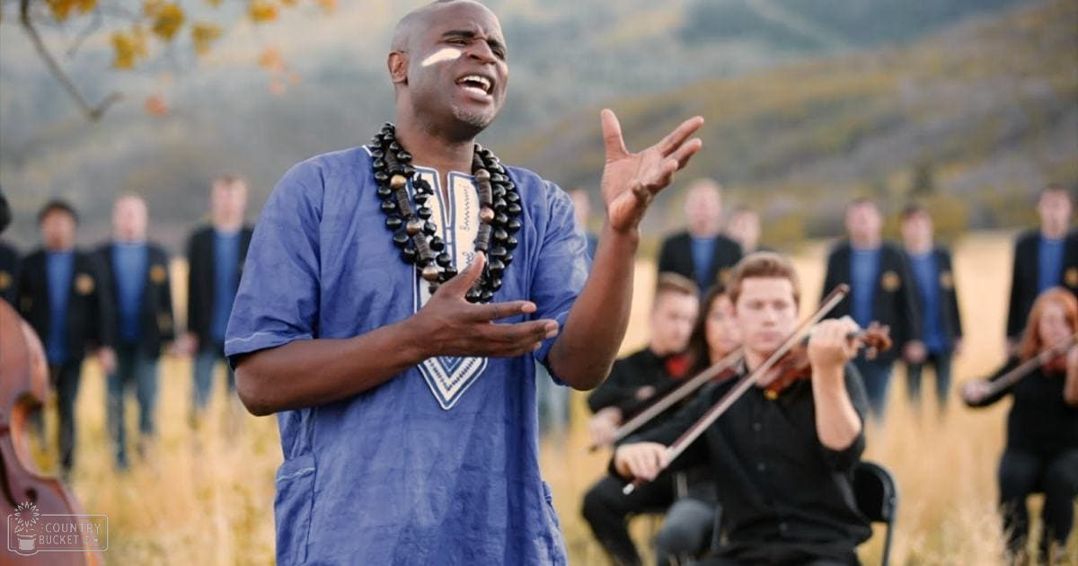 Adult male singer performing outdoors in a field surrounded by musicians, with dry grass and blurred hills in the background suggesting an autumnal setting.