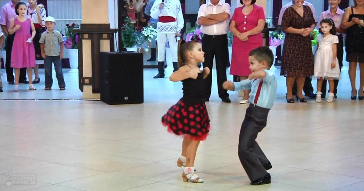 Young children dancing at an indoor social gathering with adults dressed formally, surrounded by decorative plants and flowers under artificial lighting.