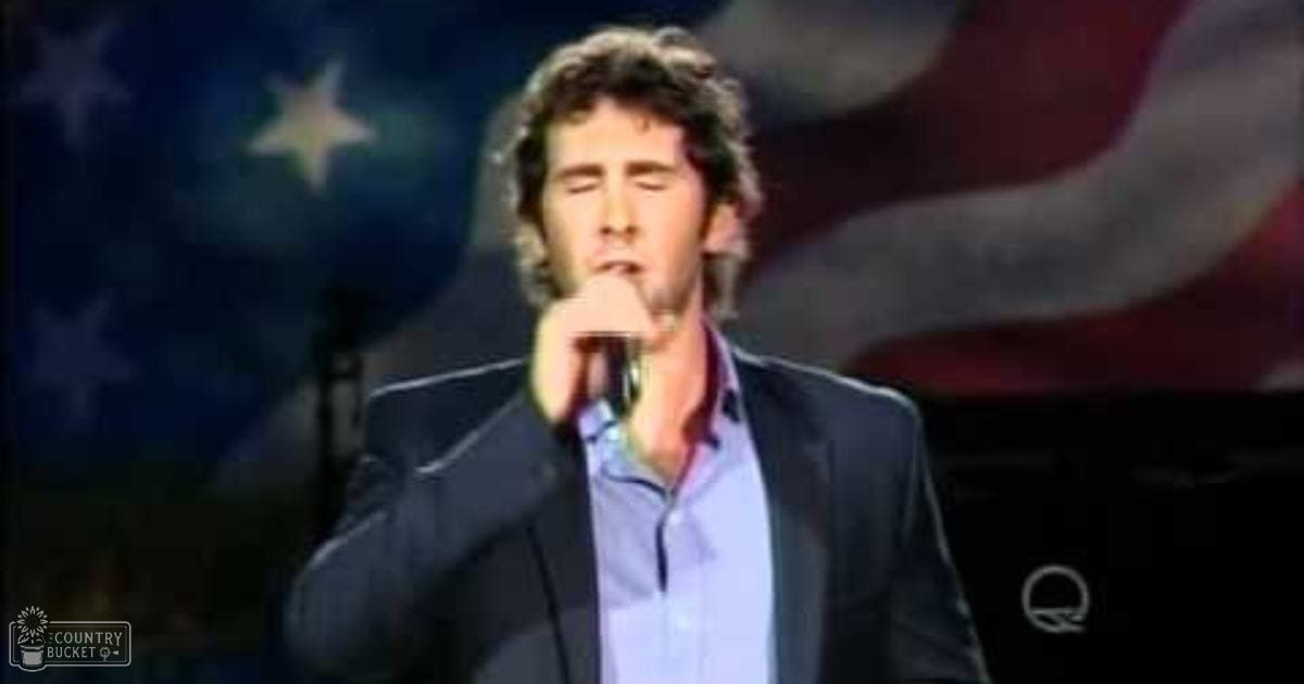 A young adult male performing on stage with an American flag backdrop, wearing a dark blazer over a light blue button-up shirt.