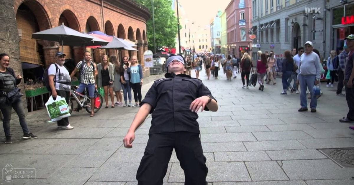 An adult female blindfolded performer in a black uniform stands in an urban city square, with onlookers and shopping activity around her. Several open umbrellas are nearby, and a sign is partially visible behind the performer.