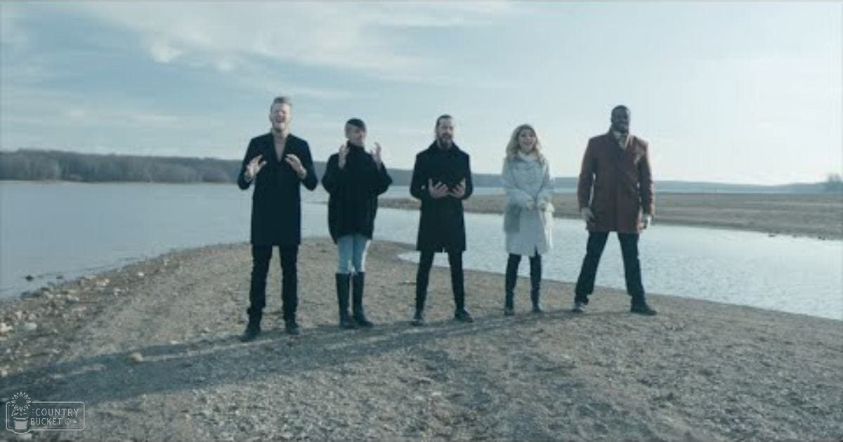 A group of five young adults dressed in dark coats stand outdoors near a body of water with vegetation and partly cloudy sky visible in the background.