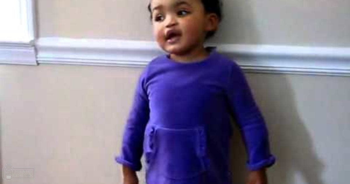 A toddler wearing a long-sleeved purple dress indoors with natural light coming from the window area.