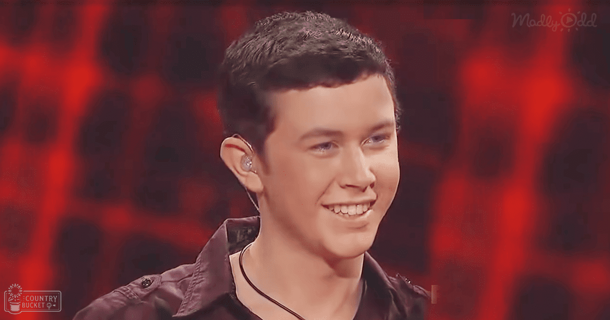 A young adult male wearing a dark brown jacket with visible buttons on shoulder and collared shirt underneath, performing indoors under red stage lighting.