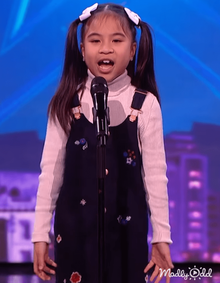 A young female child in a turtleneck sweater with floral overalls performs on stage at an event, set against blurred cityscape imagery under blue lighting.