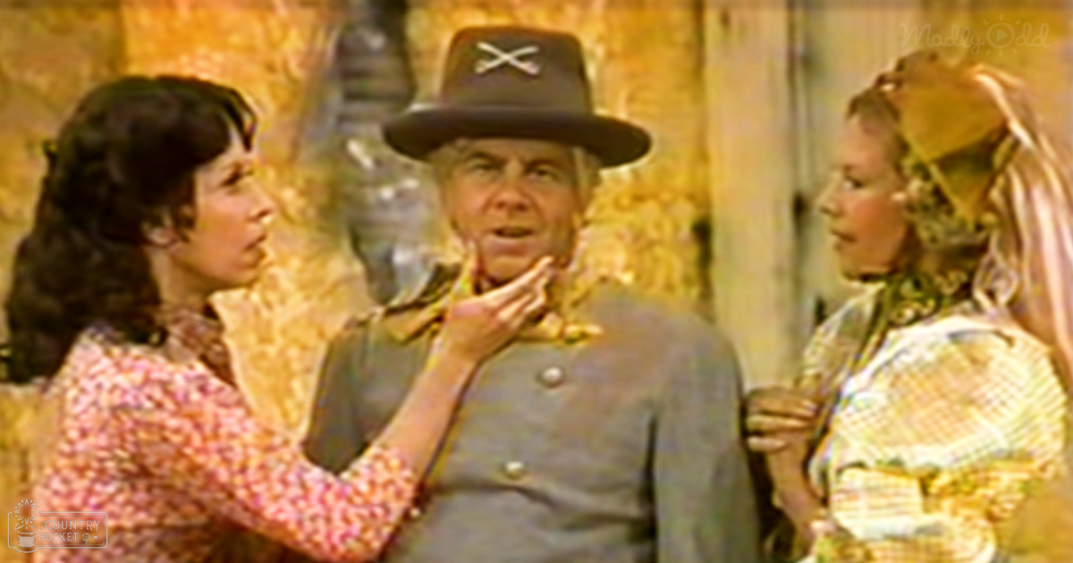 An adult male singer in a cowboy hat with crossed swords, surrounded by two adult females wearing floral blouses indoors.