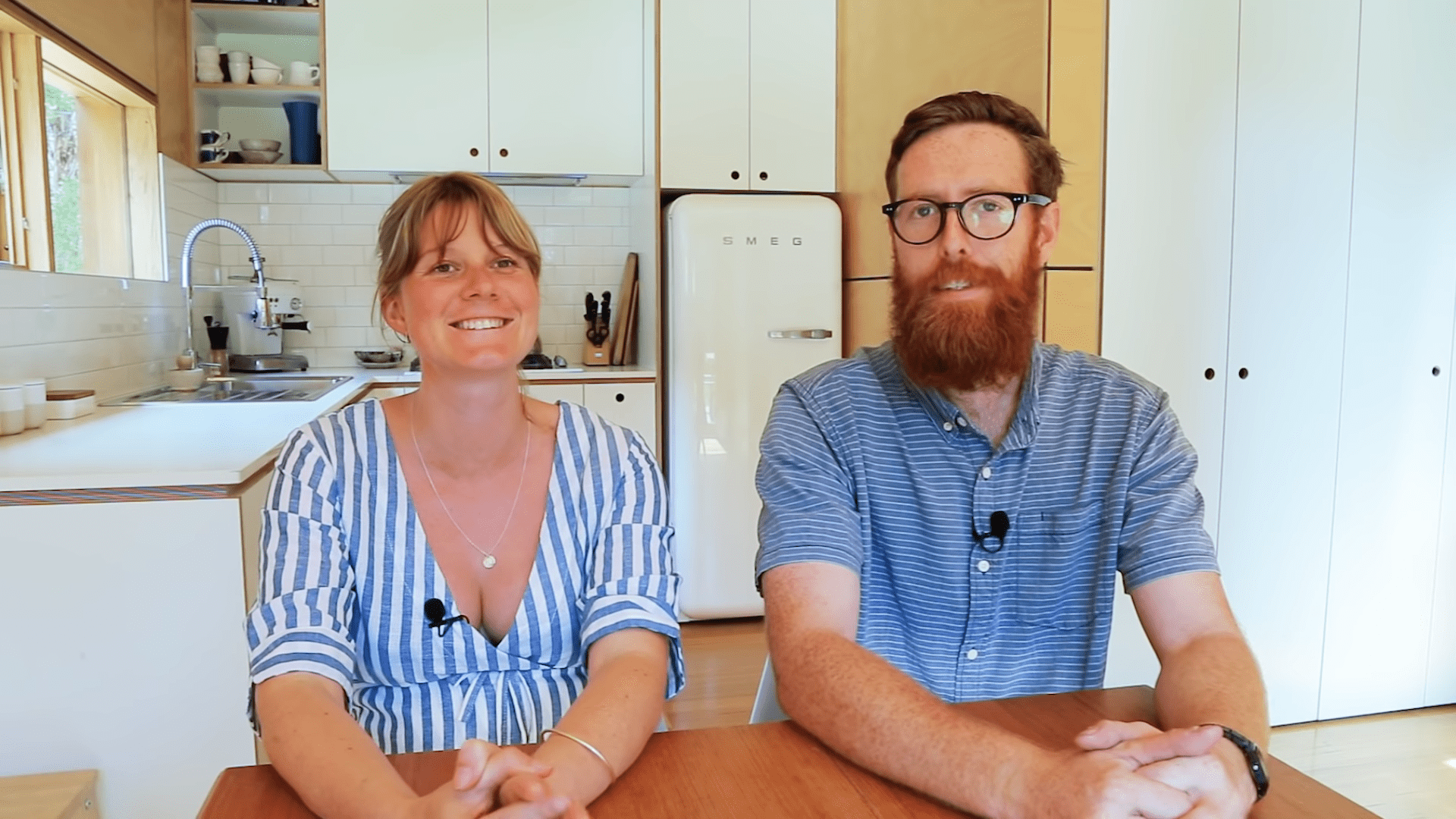 A young adult to middle-aged couple in a modern kitchen setting. The woman wears a low-cut, blue and white striped blouse with cotton fabric, while the man dons a button-down collar shirt of similar style.