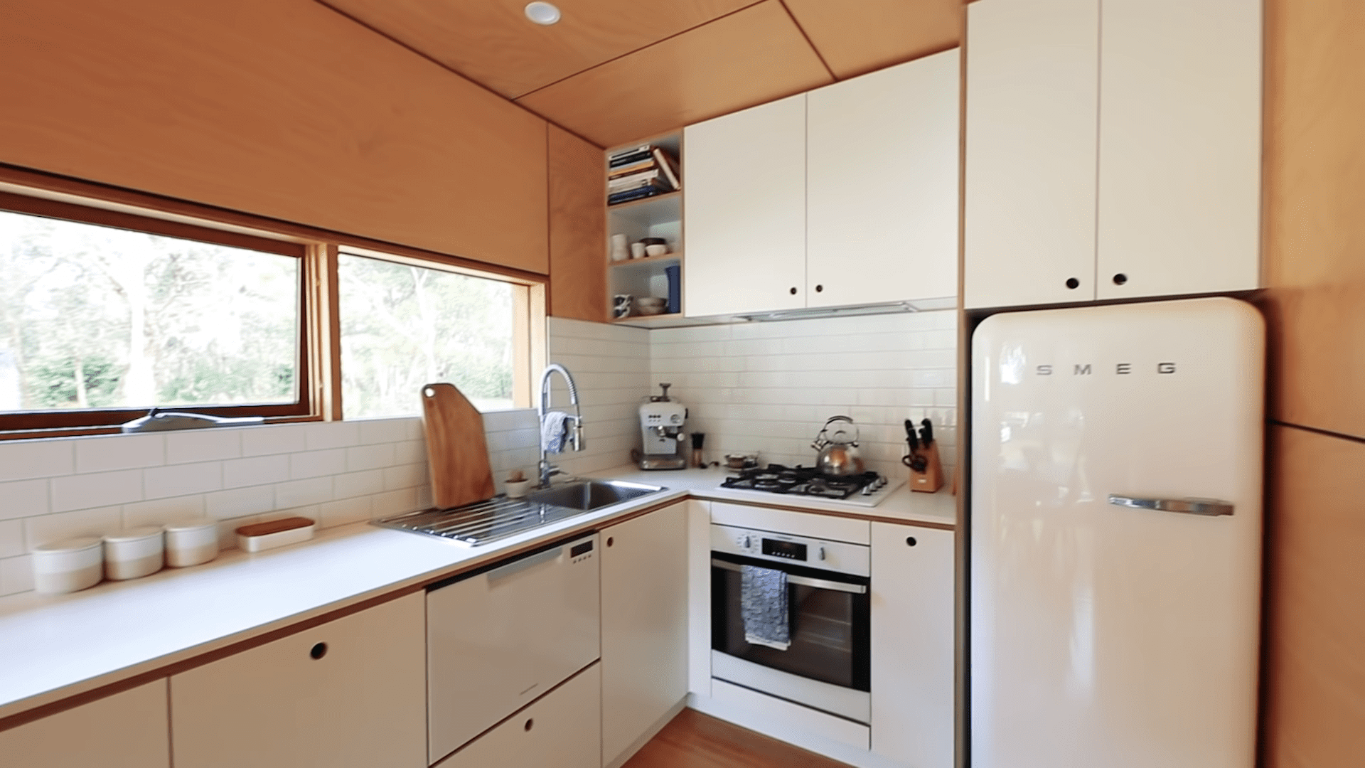 A residential kitchen interior with white SMEG refrigerator, white cabinetry, a sink with a faucet, wooden cutting board on the counter, stove with kettle and utensils, shelves containing books and bowls, natural light from window, spring or summer season, daytime setting.