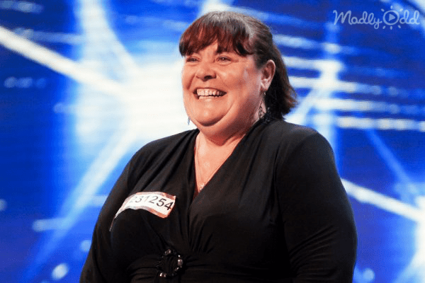 A middle-aged woman in a relaxed fit, modern casual or performance attire with long sleeves and a V-neck, wearing the number "731254" pinned on her left arm, standing against a backdrop labeled 'Madly Odd' under blue stage lighting during what appears to be a talent show.