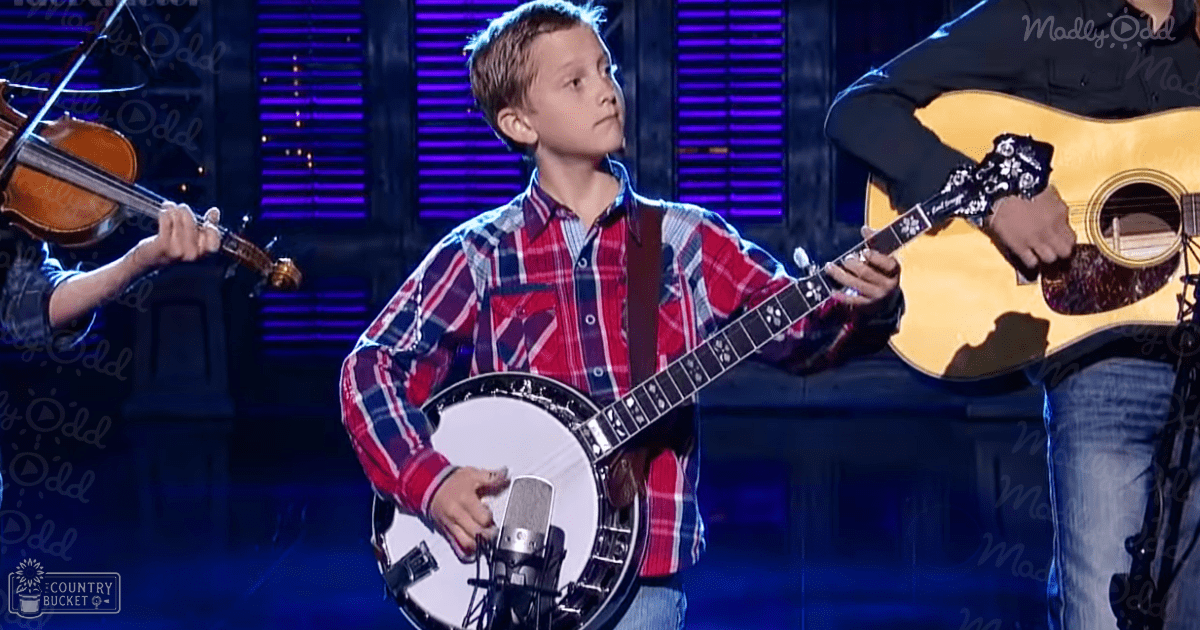 A young boy in a plaid red and blue long-sleeved shirt, blue jeans, performs with musical instruments on an indoor stage under purple lighting.