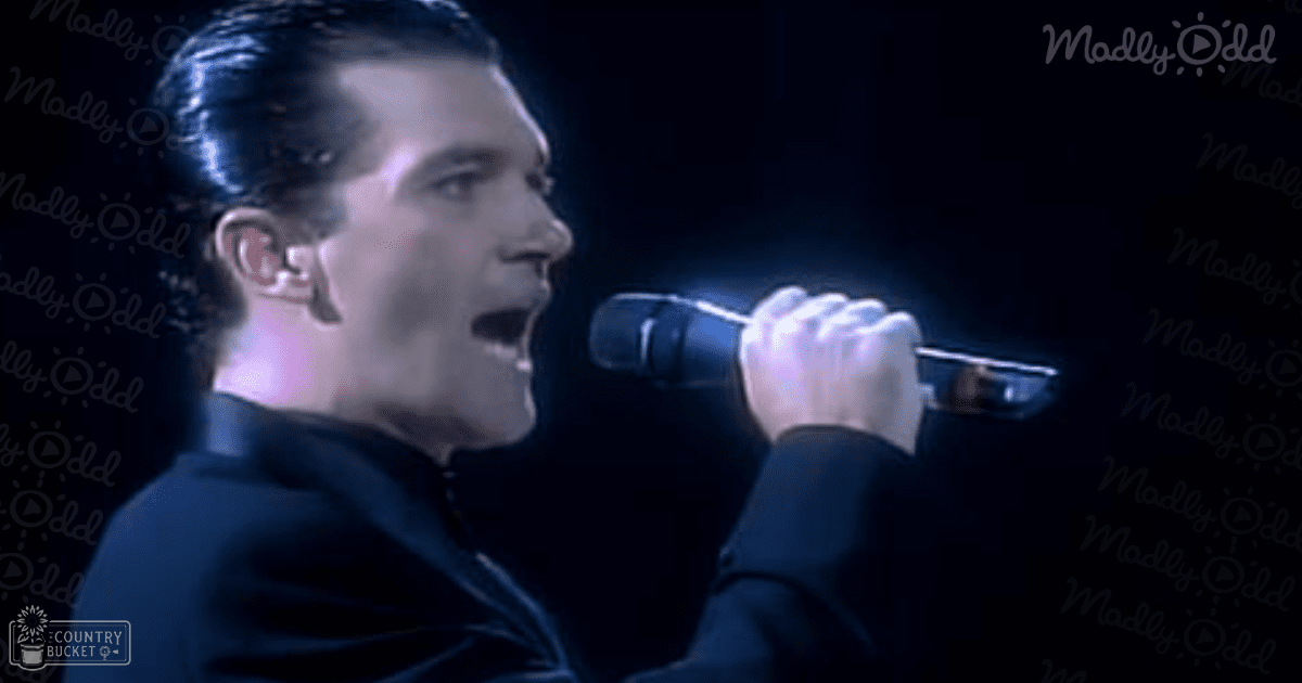 A young adult male performer in a dark zip-up jacket sings into a professional stage microphone against a dark background with 'Madly Odd' text overlay.