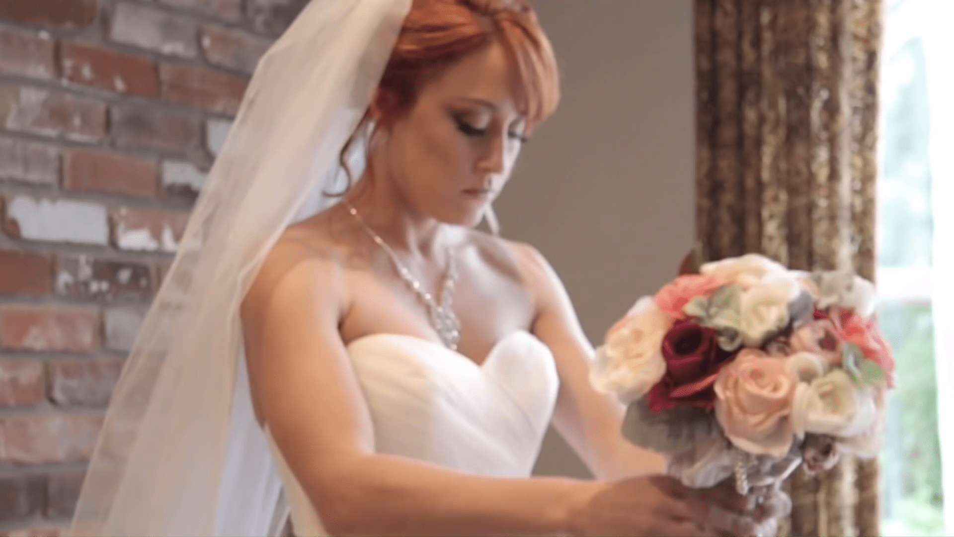 A bride wearing a strapless wedding dress with a fitted bodice and full skirt, holding a bouquet of flowers in an indoor setting with exposed brick walls and patterned curtains.