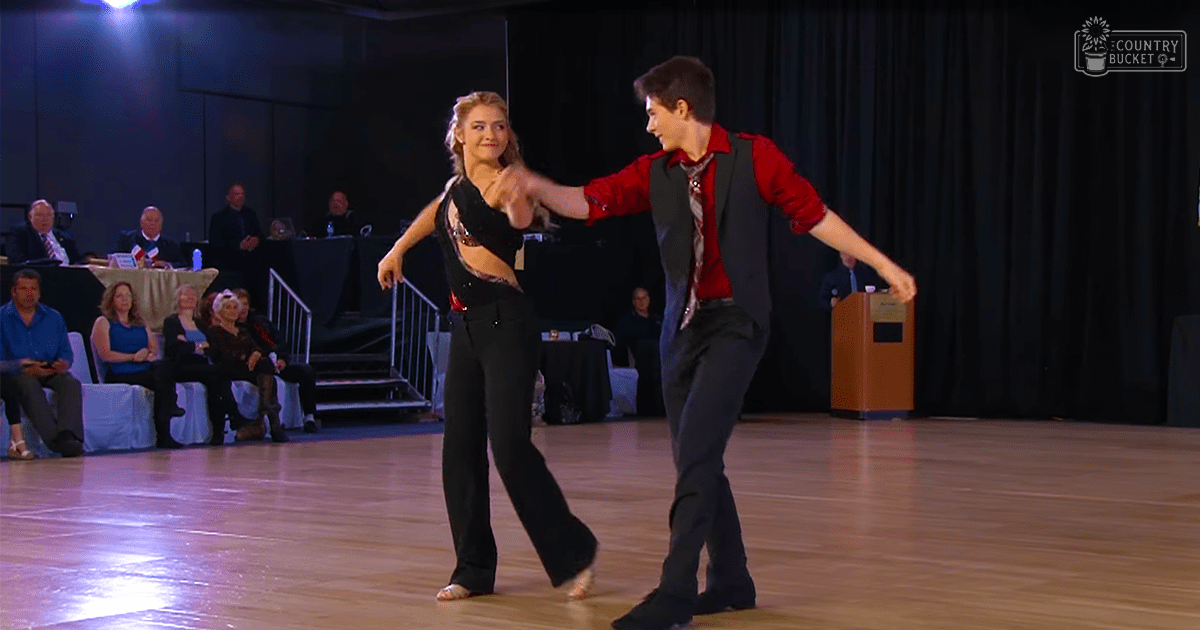 A dance couple performing at a ballroom event with an audience of spectators seated in the background. The setting is indoors, likely in an auditorium or community center, with artificial lighting suggesting evening or night.
