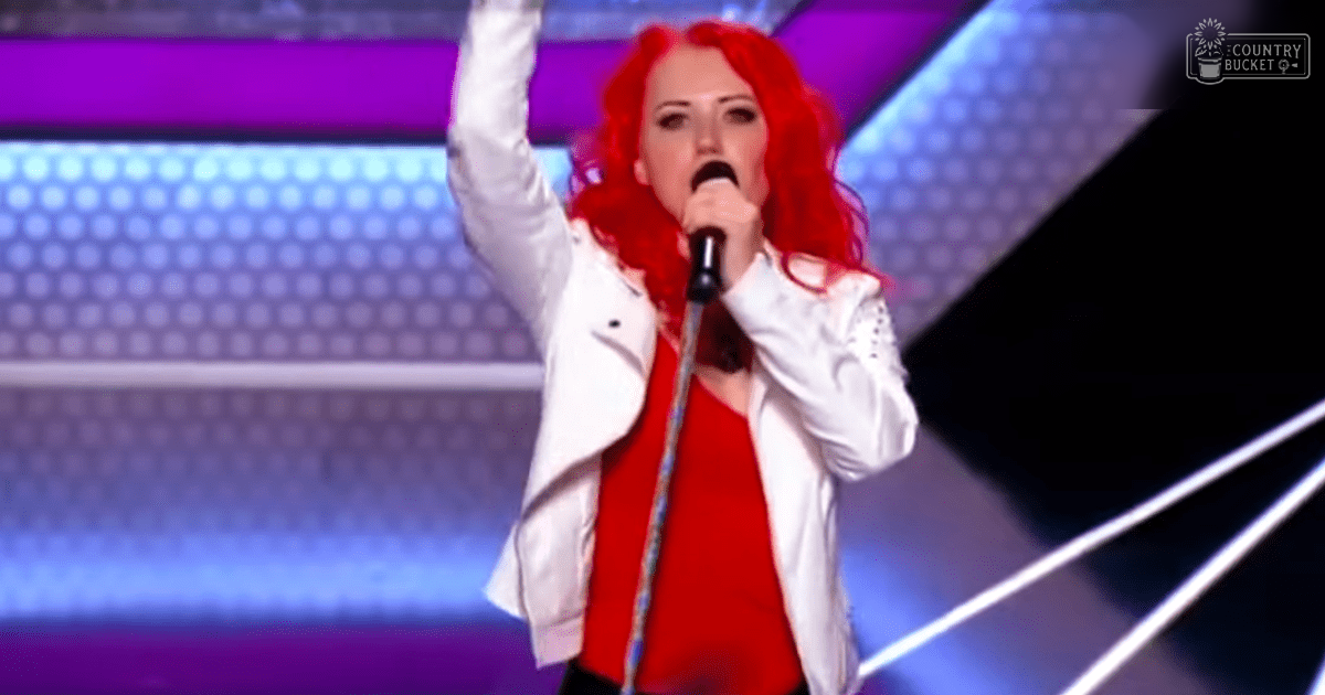 A young adult female in a white jacket with black detailing and red inner garment performs on stage at an X Factor audition, holding a microphone under bright stage lighting.