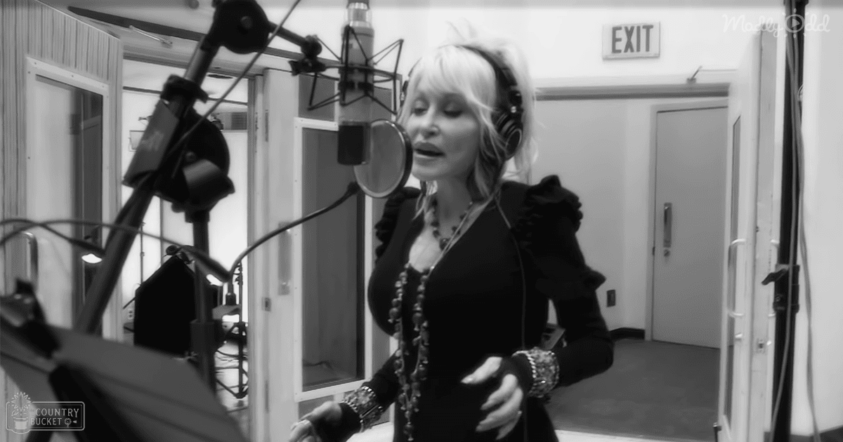 Adult female with long blonde hair, wearing headphones, a black ruffled-sleeve top, necklace, bracelets, and watch in an indoor recording studio setting.