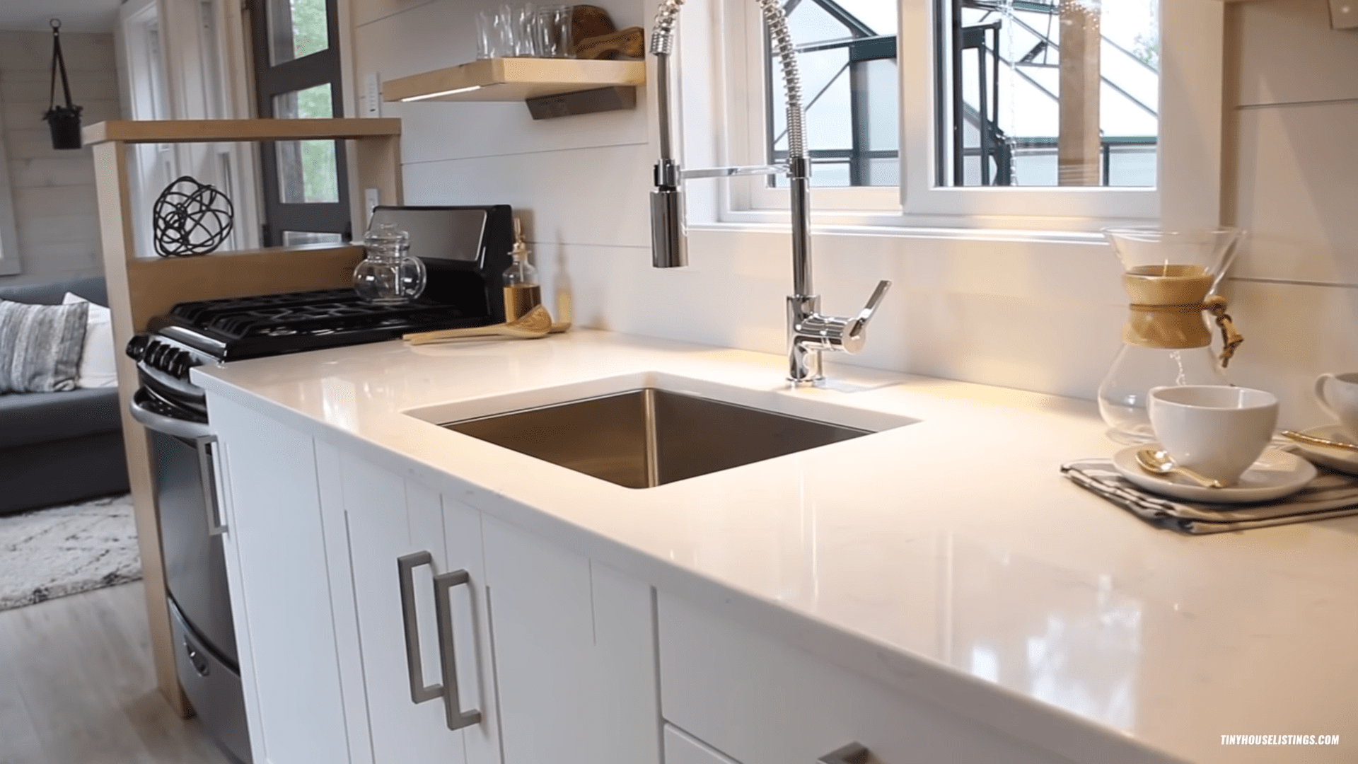 A modern kitchen interior with white countertops, black stovetop oven, stainless steel sink, neatly arranged glassware, and natural daylight streaming through windows.