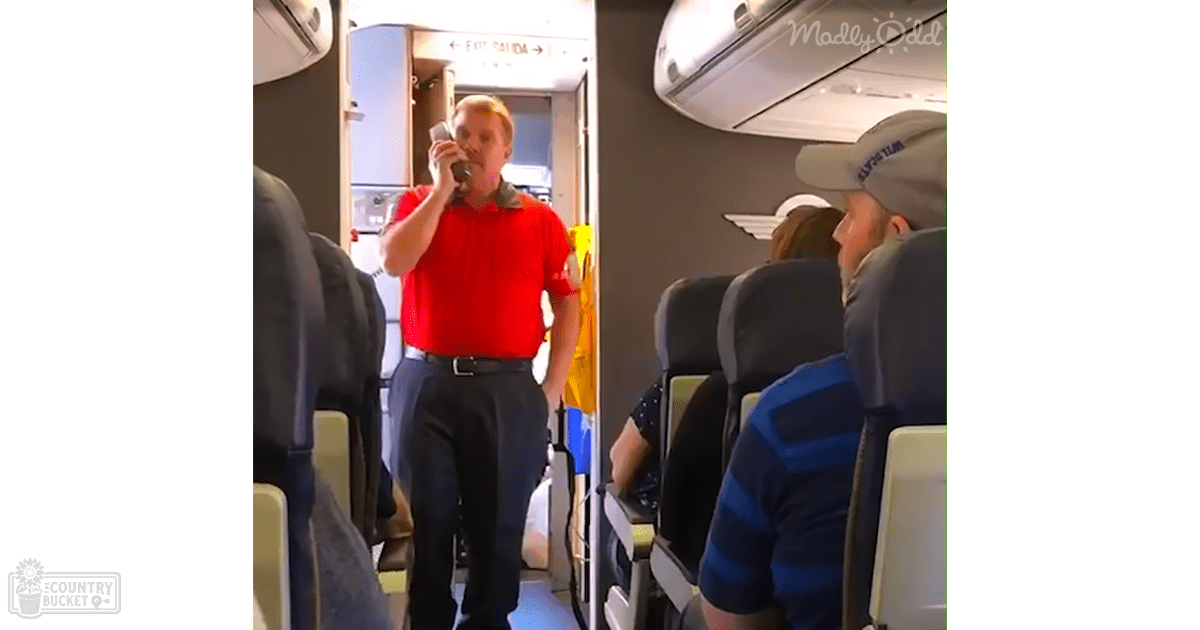 Inside an airplane cabin with a male flight attendant wearing a red polo shirt and dark pants, holding a microphone while speaking to passengers. The setting includes overhead compartments, seats, and safety vests hanging on the wall.