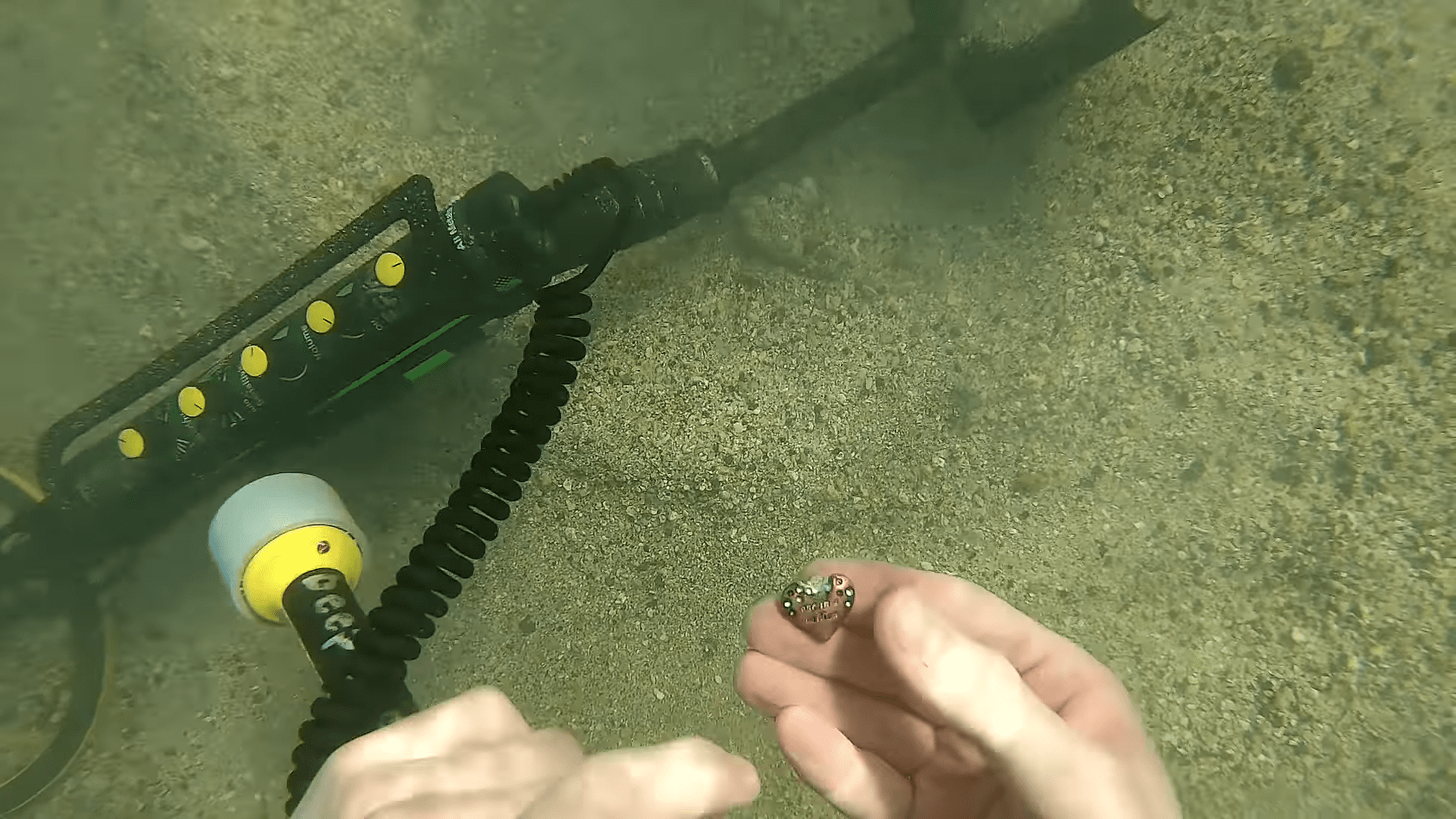 Hands holding small object near handheld metal detector in underwater riverbed setting.