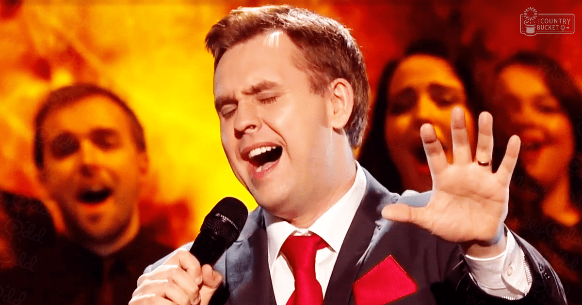 An adult male singer in a dark suit with a white shirt, red tie, and pocket square, performing on an indoor stage under warm orange lighting.