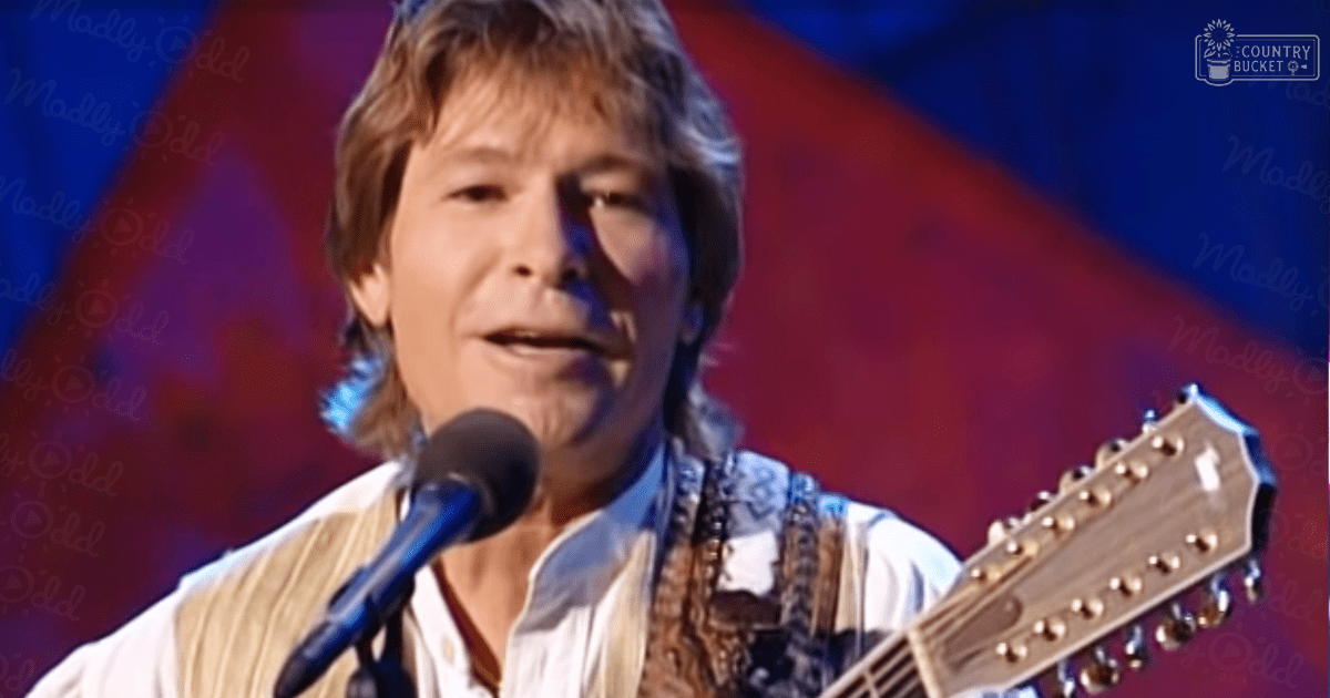 A middle-aged male performer wearing a white button-down shirt with vertical stripes and a vest, holding an acoustic guitar on stage under red and blue lighting during what appears to be an indoor concert.