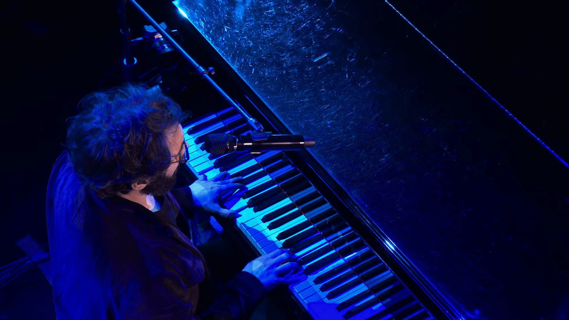 Adult male musician in a relaxed fit long-sleeved shirt playing piano at an indoor concert with stage lighting and professional audio equipment.