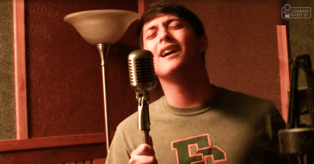 A young adult male singer wearing a green t-shirt with red text, holding a vintage-style microphone in an indoor setting with warm lighting and textured wallpaper.