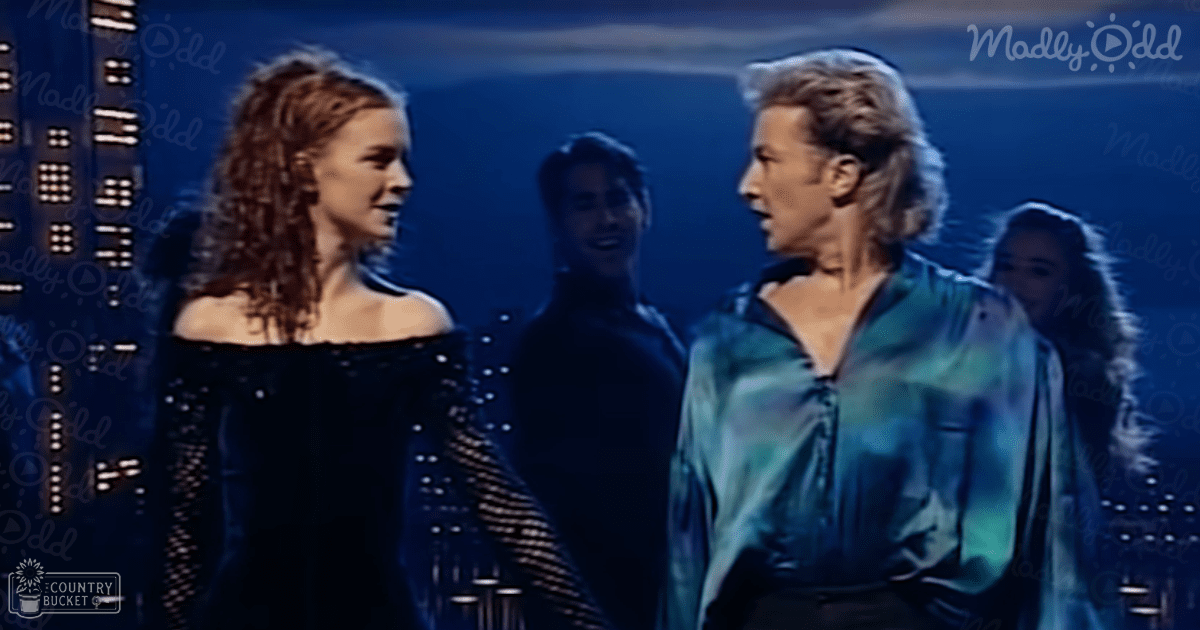 A young adult female in an off-the-shoulder, patterned black dress stands with a male companion wearing a casual teal shirt in an urban setting at night.