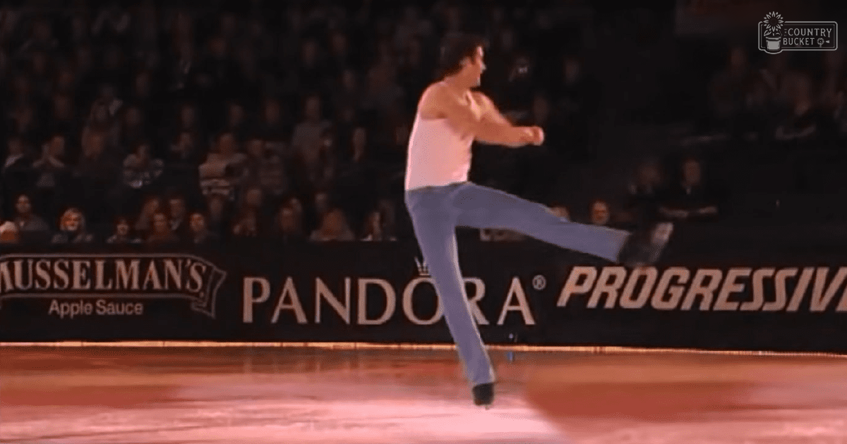Adult male figure skater performing at an indoor arena with advertisements for 'Musselman's Apple Sauce,' 'Pandora,' and 'Progressive' displayed on banners along the edge of the rink.