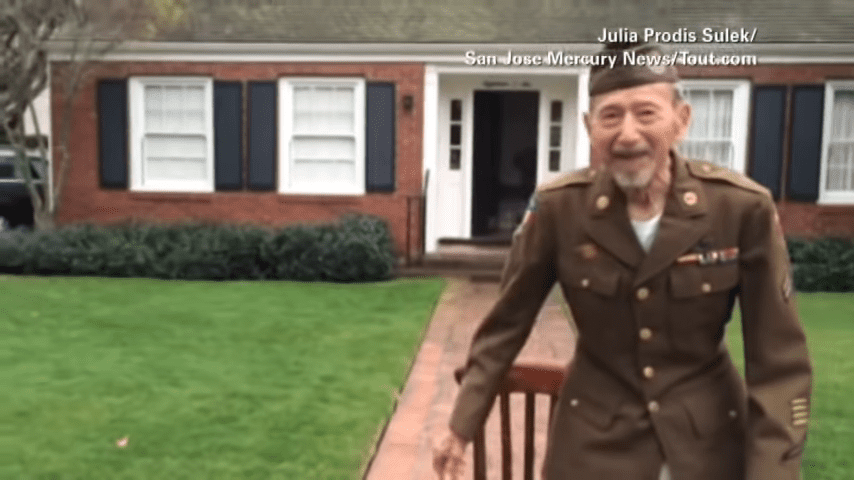 An older adult male in a military uniform stands in front of his home, surrounded by well-maintained greenery and brick architecture under soft daylight.