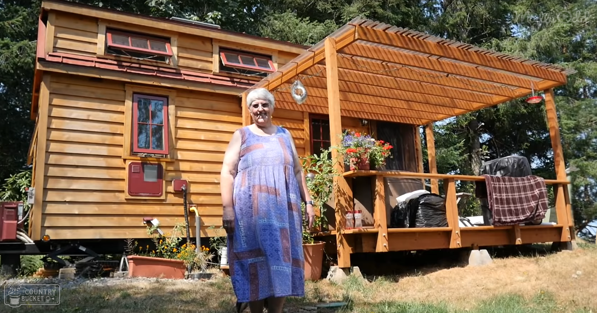 An older adult female in a loose, knee-length sleeveless dress with square patterns stands outdoors near a small wooden house with red-framed windows. The scene includes potted plants and a gravel path surrounded by greenery.