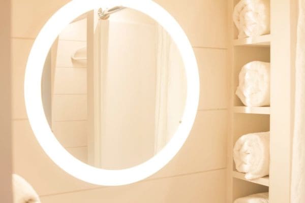 A bathroom scene with a circular mirror with bright lighting, neatly folded towels on shelves to the right, and a tiled wall suggesting a bathtub or shower area.