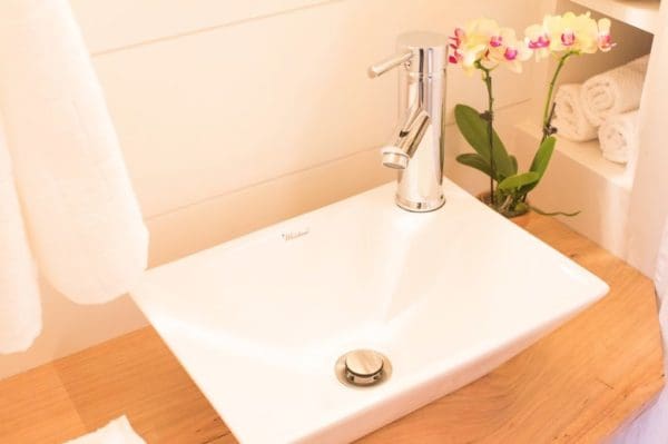 Bathroom interior with white sink, modern chrome faucet, wooden countertop, potted orchid, folded towels, natural or soft artificial lighting.