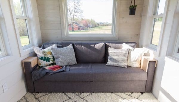 A cozy living room corner with a dark gray sofa adorned with floral and textured pillows, light-colored paneling walls, white rug under furniture, small potted plant on the wall, natural light from large windows, outdoor view of an open field or rural area with trees in spring or summer, daytime scene.
