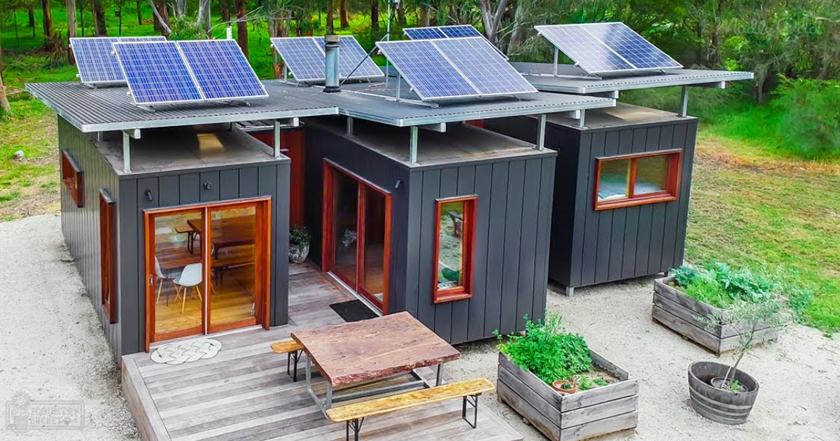 A modern house with dark gray siding and red-framed windows, with solar panels on its roof in a lush garden setting surrounded by trees and grass.