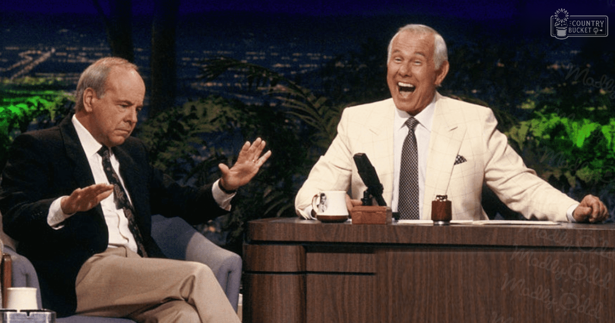 A late-night talk show set with two older adult males seated at a desk, wearing dark and light checkered suits against an out-of-focus nighttime cityscape backdrop.