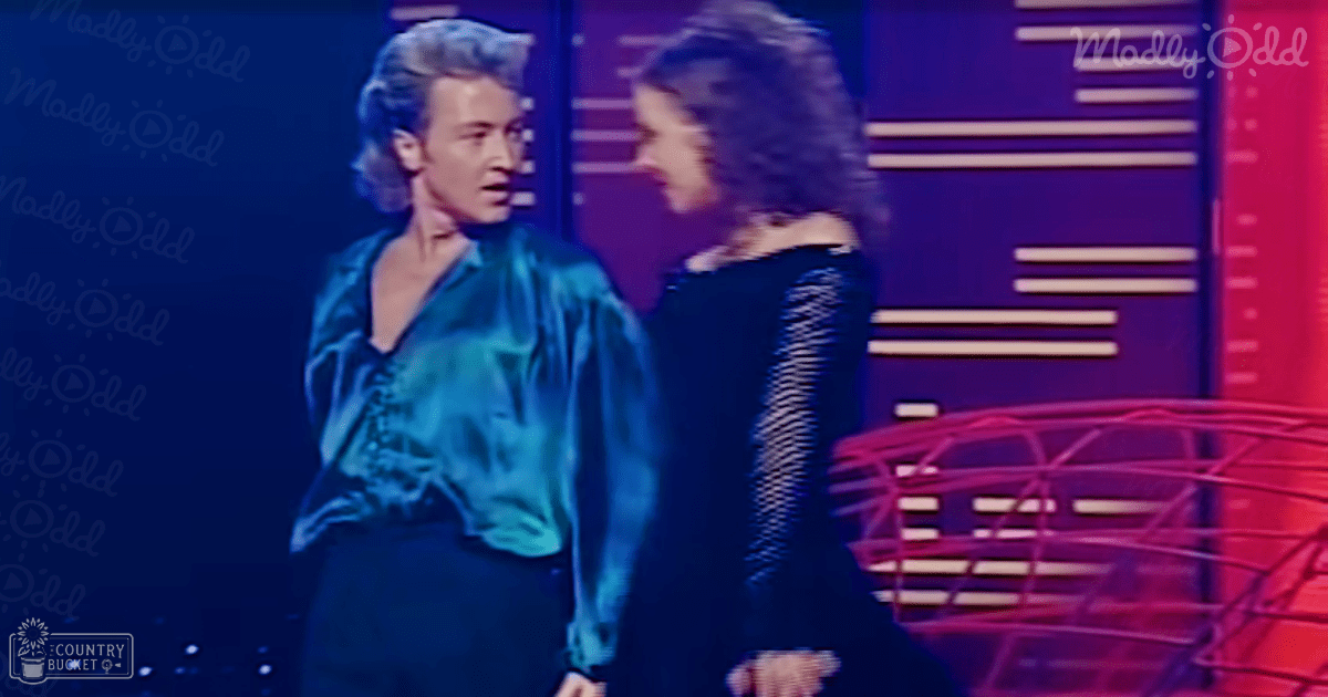 A young adult male singer wearing a teal shirt with patterned design and dark pants stands beside another young adult female performer in a dark textured top on an indoor stage set against purple-blue vertical panel backdrop at nighttime.