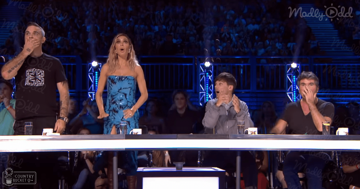 A talent show scene with contestants at a podium; an adult male singer in a black t-shirt with white text stands leftmost, a female contestant in a blue patterned dress is second from the left, and two more adults appear surprised or shocked. Stage lights illuminate the judges' panel.