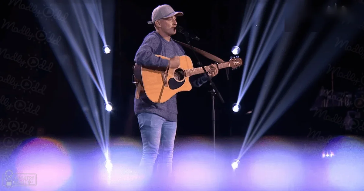 An adult male singer in a dark blue sweater and gray baseball cap performing on an indoor concert stage with bright white lighting and purple fog.