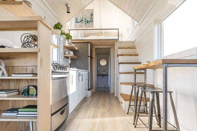 Cozy kitchen in a tiny house with wooden shelves, stainless steel appliances, and framed wall art. Natural light fills the space during midday.