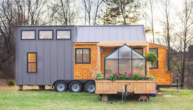 A tiny house with a gray exterior, wooden accents, large windows, and a greenhouse filled with flowers sits on wheels in an outdoor setting surrounded by trees without leaves.
